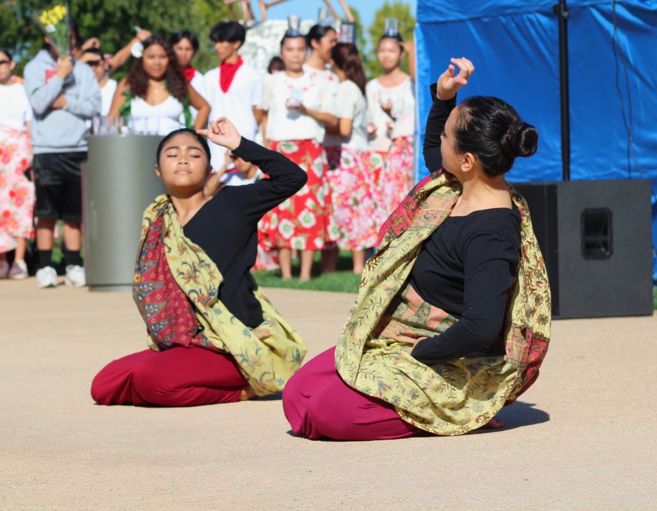 Two dancers performing in coordinated maroon, black, green, and gold costumes at the outdoor festival.