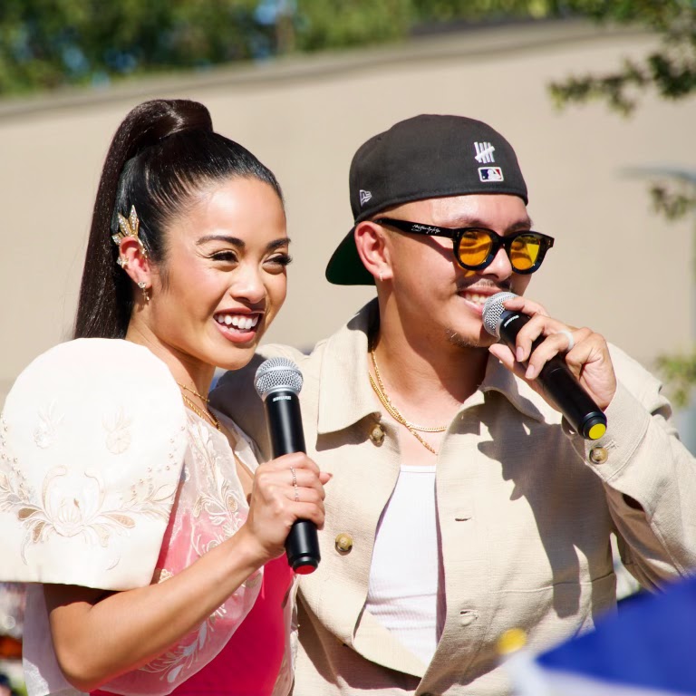 Two smiling festival presenters holding microphones on an outdoor stage.