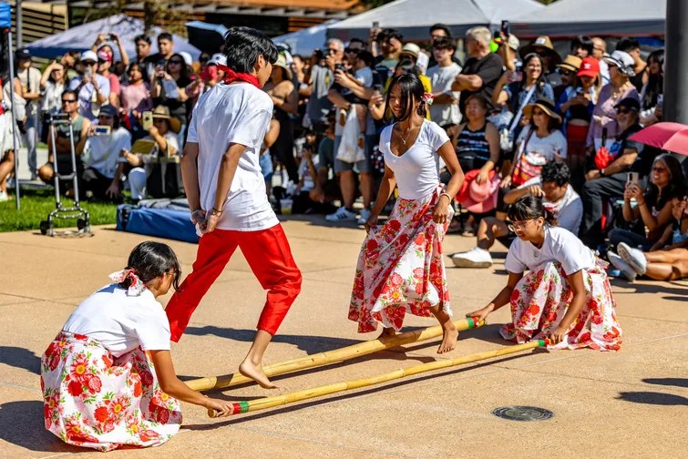 Young dancers performing tinikling with bamboo poles as a festival crowd watches.